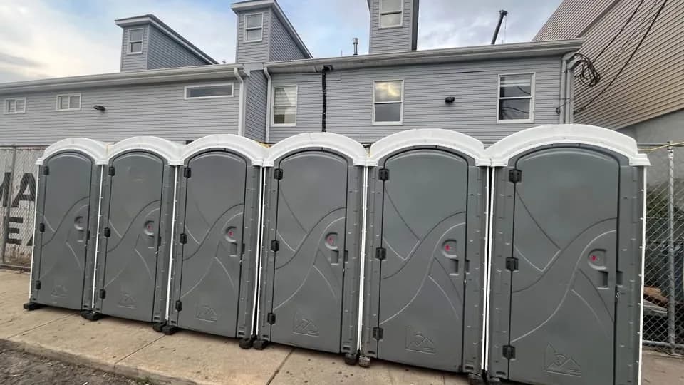 Six portable restrooms lined up along a paved lot beside a building