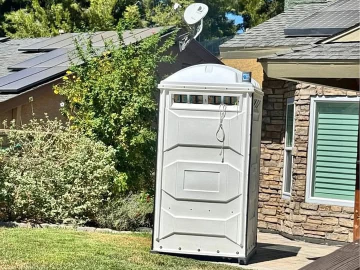 Tall vented portable restroom standing on a lawn beside a house